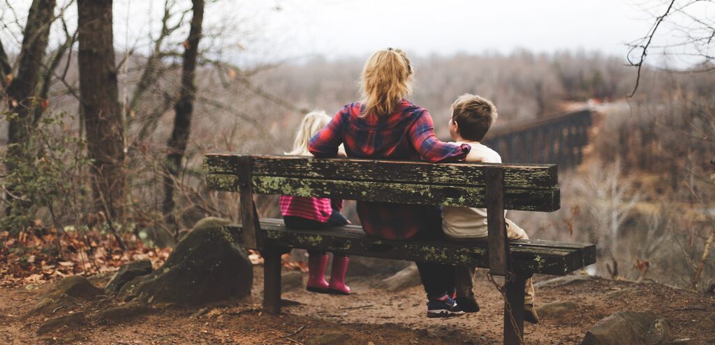 Una madre sentada en un banco del bosque con sus dos hijos, de espaldas