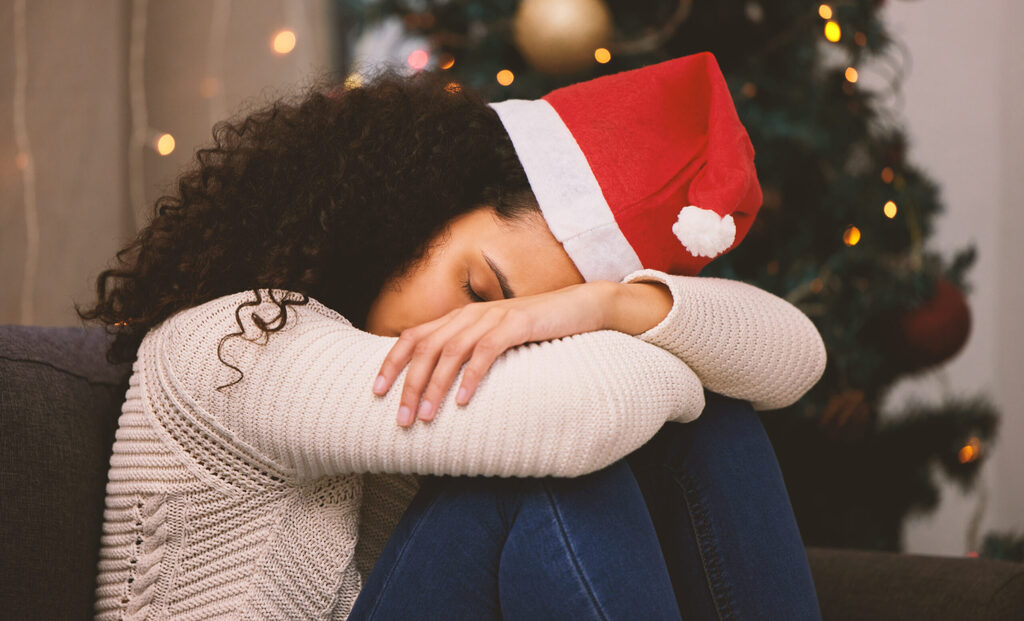 Una mujer con gorro de Papá Noel apoya la cabeza en las rodillas junto al árbol
